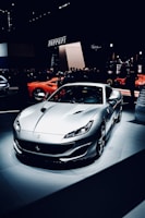 A sleek, silver sports car is prominently displayed under spotlights, surrounded by a crowd in a dimly lit showroom. The background features branding or signage related to the manufacturer.