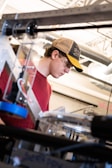 A person wearing a red shirt and a baseball cap is focused on working with equipment in an industrial or workshop setting. Safety glasses are worn, and various mechanical components and tools are visible in the foreground and background, indicating a technical or engineering environment.
