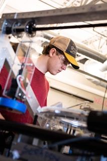 A person wearing a red shirt and a baseball cap is focused on working with equipment in an industrial or workshop setting. Safety glasses are worn, and various mechanical components and tools are visible in the foreground and background, indicating a technical or engineering environment.