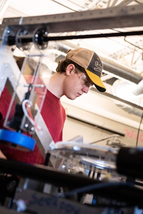 A person wearing a red shirt and a baseball cap is focused on working with equipment in an industrial or workshop setting. Safety glasses are worn, and various mechanical components and tools are visible in the foreground and background, indicating a technical or engineering environment.