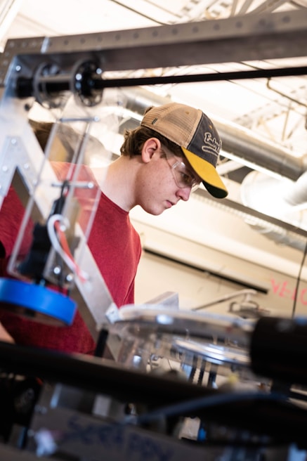 A person wearing a red shirt and a baseball cap is focused on working with equipment in an industrial or workshop setting. Safety glasses are worn, and various mechanical components and tools are visible in the foreground and background, indicating a technical or engineering environment.