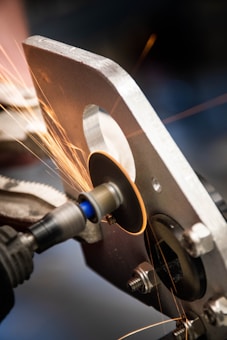 A metal grinding process where sparks are flying as a circular grinding tool is working on a piece of metal. The scene appears to be in a workshop setting with detailed focus on the machine and the metal piece.