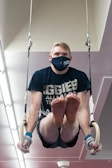 A person is performing a gymnastics routine on rings. They are wearing a navy mask with 'Aggie Strong' printed on it and a matching navy shirt. The individual is suspended mid-air, gripping the rings firmly, demonstrating strength and focus.