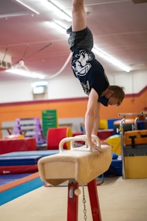 A person is performing a handstand on a pommel horse in a gymnasium, with various gym equipment visible in the background. The individual is wearing a black t-shirt and shorts, and their face is partially covered by a mask.