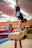 A person is performing a handstand on a pommel horse in a gymnasium, with various gym equipment visible in the background. The individual is wearing a black t-shirt and shorts, and their face is partially covered by a mask.