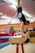 A person is performing a handstand on a pommel horse in a gymnasium, with various gym equipment visible in the background. The individual is wearing a black t-shirt and shorts, and their face is partially covered by a mask.