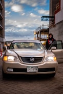 A silver Acura car is parked in an urban setting near building constructions. The front lights on the vehicle are turned on, and there is a visible license plate from Colorado. A person wearing a dark hoodie stands beside the open passenger door. The scene is set under a partly cloudy sky, with construction scaffolding visible in the background.