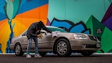A person wearing a black jacket and jeans is cleaning a beige sedan with a cloth next to a colorful mural. The mural features bold geometric shapes with bright colors, including green, blue, orange, and black. The scene is set on a street with a slightly overcast look.