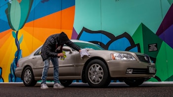 A person wearing a black jacket and jeans is cleaning a beige sedan with a cloth next to a colorful mural. The mural features bold geometric shapes with bright colors, including green, blue, orange, and black. The scene is set on a street with a slightly overcast look.