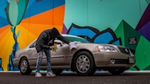 A person wearing a black jacket and jeans is cleaning a beige sedan with a cloth next to a colorful mural. The mural features bold geometric shapes with bright colors, including green, blue, orange, and black. The scene is set on a street with a slightly overcast look.