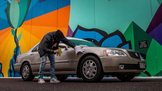A person wearing a black jacket and jeans is cleaning a beige sedan with a cloth next to a colorful mural. The mural features bold geometric shapes with bright colors, including green, blue, orange, and black. The scene is set on a street with a slightly overcast look.