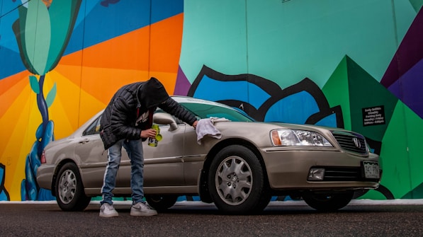 A person wearing a black jacket and jeans is cleaning a beige sedan with a cloth next to a colorful mural. The mural features bold geometric shapes with bright colors, including green, blue, orange, and black. The scene is set on a street with a slightly overcast look.
