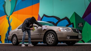 A person wearing a black jacket and jeans is cleaning a beige sedan with a cloth next to a colorful mural. The mural features bold geometric shapes with bright colors, including green, blue, orange, and black. The scene is set on a street with a slightly overcast look.