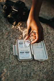 A hand is holding a Samsung 128GB Pro memory card above an open case containing several other memory cards. In the background, a Canon camera is placed on a textured surface.