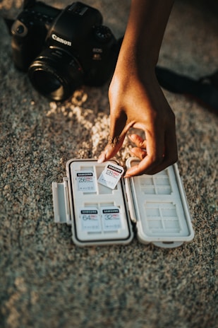A hand is holding a Samsung 128GB Pro memory card above an open case containing several other memory cards. In the background, a Canon camera is placed on a textured surface.
