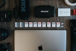The image contains a collection of technology-related objects arranged on a wooden surface. A silver laptop with an Apple logo is prominently positioned at the bottom center. Above the laptop, there is a row of various SD cards, each with different storage capacities and brands. Surrounding these are other tech accessories including a black microphone with 'RØDE' branding, a Pelican hard case, a silver Samsung portable SSD, a Sony camera battery, photographic lenses, and rolls of tape.