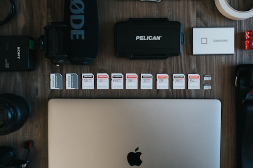 The image contains a collection of technology-related objects arranged on a wooden surface. A silver laptop with an Apple logo is prominently positioned at the bottom center. Above the laptop, there is a row of various SD cards, each with different storage capacities and brands. Surrounding these are other tech accessories including a black microphone with 'RØDE' branding, a Pelican hard case, a silver Samsung portable SSD, a Sony camera battery, photographic lenses, and rolls of tape.