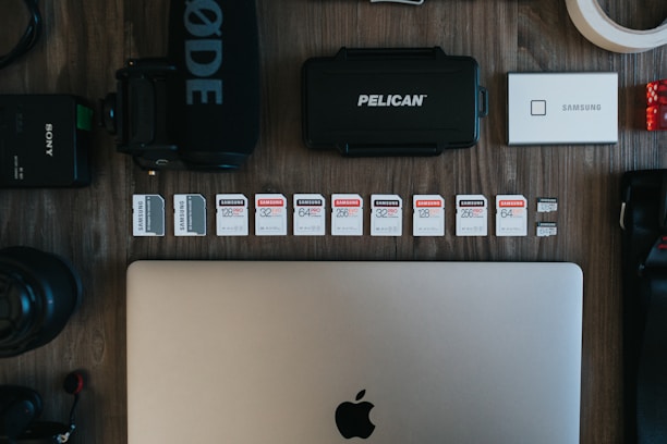 The image contains a collection of technology-related objects arranged on a wooden surface. A silver laptop with an Apple logo is prominently positioned at the bottom center. Above the laptop, there is a row of various SD cards, each with different storage capacities and brands. Surrounding these are other tech accessories including a black microphone with 'RØDE' branding, a Pelican hard case, a silver Samsung portable SSD, a Sony camera battery, photographic lenses, and rolls of tape.