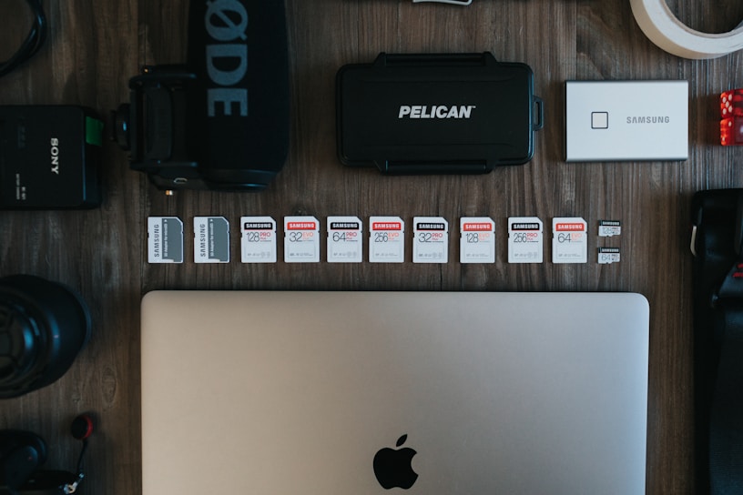 The image contains a collection of technology-related objects arranged on a wooden surface. A silver laptop with an Apple logo is prominently positioned at the bottom center. Above the laptop, there is a row of various SD cards, each with different storage capacities and brands. Surrounding these are other tech accessories including a black microphone with 'RØDE' branding, a Pelican hard case, a silver Samsung portable SSD, a Sony camera battery, photographic lenses, and rolls of tape.