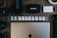 The image contains a collection of technology-related objects arranged on a wooden surface. A silver laptop with an Apple logo is prominently positioned at the bottom center. Above the laptop, there is a row of various SD cards, each with different storage capacities and brands. Surrounding these are other tech accessories including a black microphone with 'RØDE' branding, a Pelican hard case, a silver Samsung portable SSD, a Sony camera battery, photographic lenses, and rolls of tape.