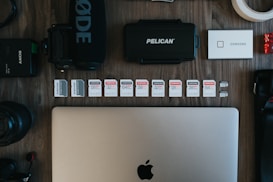 The image contains a collection of technology-related objects arranged on a wooden surface. A silver laptop with an Apple logo is prominently positioned at the bottom center. Above the laptop, there is a row of various SD cards, each with different storage capacities and brands. Surrounding these are other tech accessories including a black microphone with 'RØDE' branding, a Pelican hard case, a silver Samsung portable SSD, a Sony camera battery, photographic lenses, and rolls of tape.