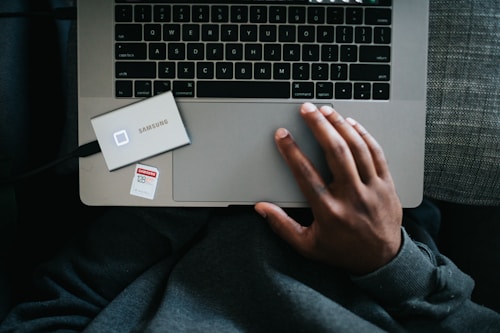 A person is using a laptop computer with their right hand on the trackpad. A Samsung external SSD is placed beside the laptop, connected via a cable. A 128GB SD card rests on the laptop palm rest area. The person is wearing a dark grey sweater.