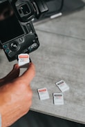 A close-up of a person inserting a 256 GB memory card into a digital camera. Several other memory cards are scattered on a gray surface nearby. The scene is focused on the camera and memory cards, highlighting their details.