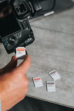 A close-up of a person inserting a 256 GB memory card into a digital camera. Several other memory cards are scattered on a gray surface nearby. The scene is focused on the camera and memory cards, highlighting their details.