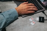 A person wearing a light denim shirt is typing on a laptop placed on a gray table. Next to the laptop, there are multiple memory cards of varying capacities and a black card reader labeled 'INSIGNIA'.