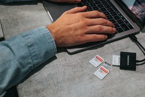A person wearing a light denim shirt is typing on a laptop placed on a gray table. Next to the laptop, there are multiple memory cards of varying capacities and a black card reader labeled 'INSIGNIA'.