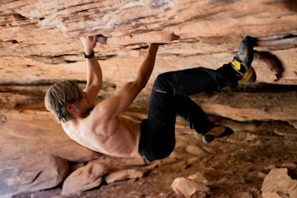 A shirtless individual with blonde hair and athletic build is engaged in rock climbing on a rocky overhang. They are using their hands and feet to grip the rough surface. The environment appears to be an arid, natural rock formation with earthy tones.