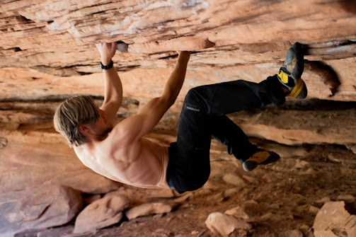A shirtless individual with blonde hair and athletic build is engaged in rock climbing on a rocky overhang. They are using their hands and feet to grip the rough surface. The environment appears to be an arid, natural rock formation with earthy tones.