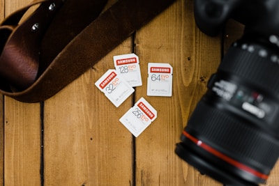 A wooden surface is arranged with four Samsung EVO Plus memory cards of varying storage capacities, alongside part of a camera lens and a brown strap.