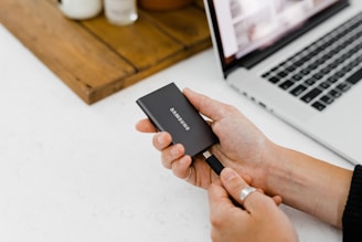 A person is holding a Samsung portable solid-state drive near a laptop. The scene is on a clean, white surface with a wooden tray holding some small containers in the background.