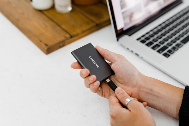 A person is holding a Samsung portable solid-state drive near a laptop. The scene is on a clean, white surface with a wooden tray holding some small containers in the background.