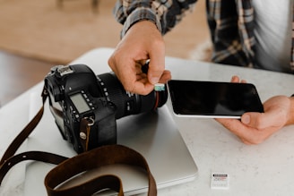 A person is holding a smartphone while inserting a memory card into it. On the table, there is a professional camera, an open laptop, and another memory card. The setting suggests a focus on photography or data management.