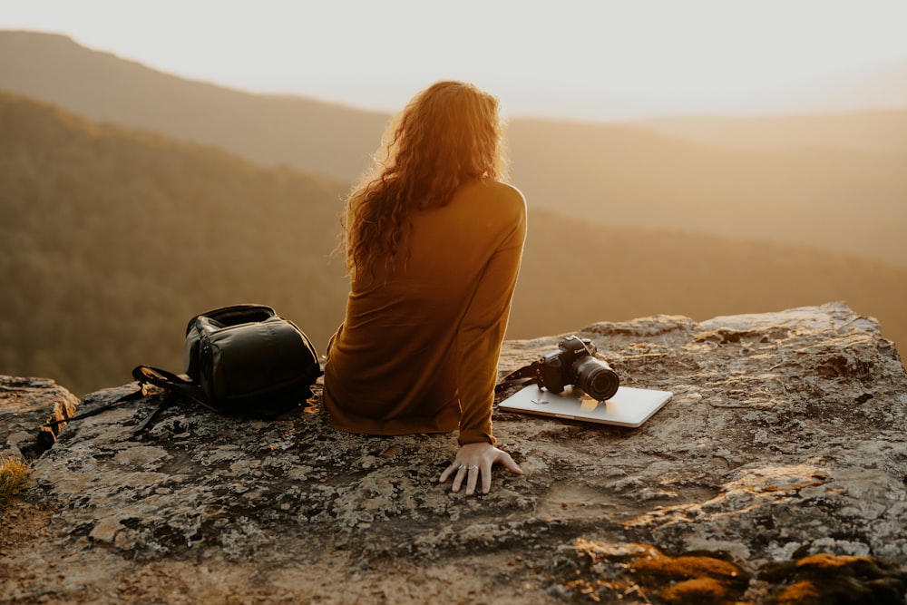 woman in brown long sleeve shirt sitting on rock near black leather bag during daytime