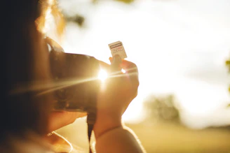 A person is holding a camera up to their face while focusing on an SD card with a 256GB capacity label. The bright sunlight creates a warm glow, causing some lens flare and casting soft light and shadows across the scene.