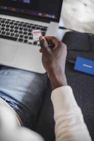 A person is holding a Samsung EVO 256GB memory card over a laptop. The scene is set on a couch, and the person is wearing a light-colored sweater and jeans. A blue external hard drive is on the couch beside the person.