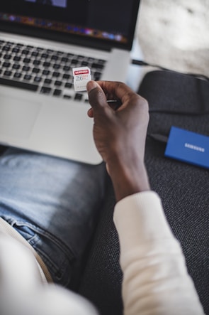 A person is holding a Samsung EVO 256GB memory card over a laptop. The scene is set on a couch, and the person is wearing a light-colored sweater and jeans. A blue external hard drive is on the couch beside the person.