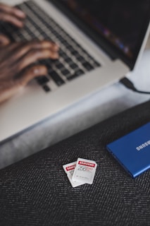 A person is typing on a laptop, with a focus on their hands on the keyboard. In the foreground, there are two Samsung memory cards and a blue Samsung external hard drive placed on a textured surface.