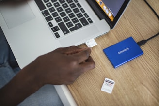 A person is inserting an SD card into a slot on a laptop. A blue external hard drive is connected to the laptop with a cable, and another SD card is lying on the wooden table nearby.