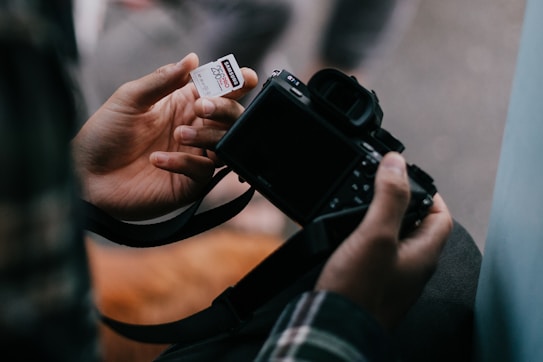 A person is holding a digital camera in one hand while placing a SanDisk memory card in the other. The camera appears to be ready for use, suggesting a photography or data storage theme.