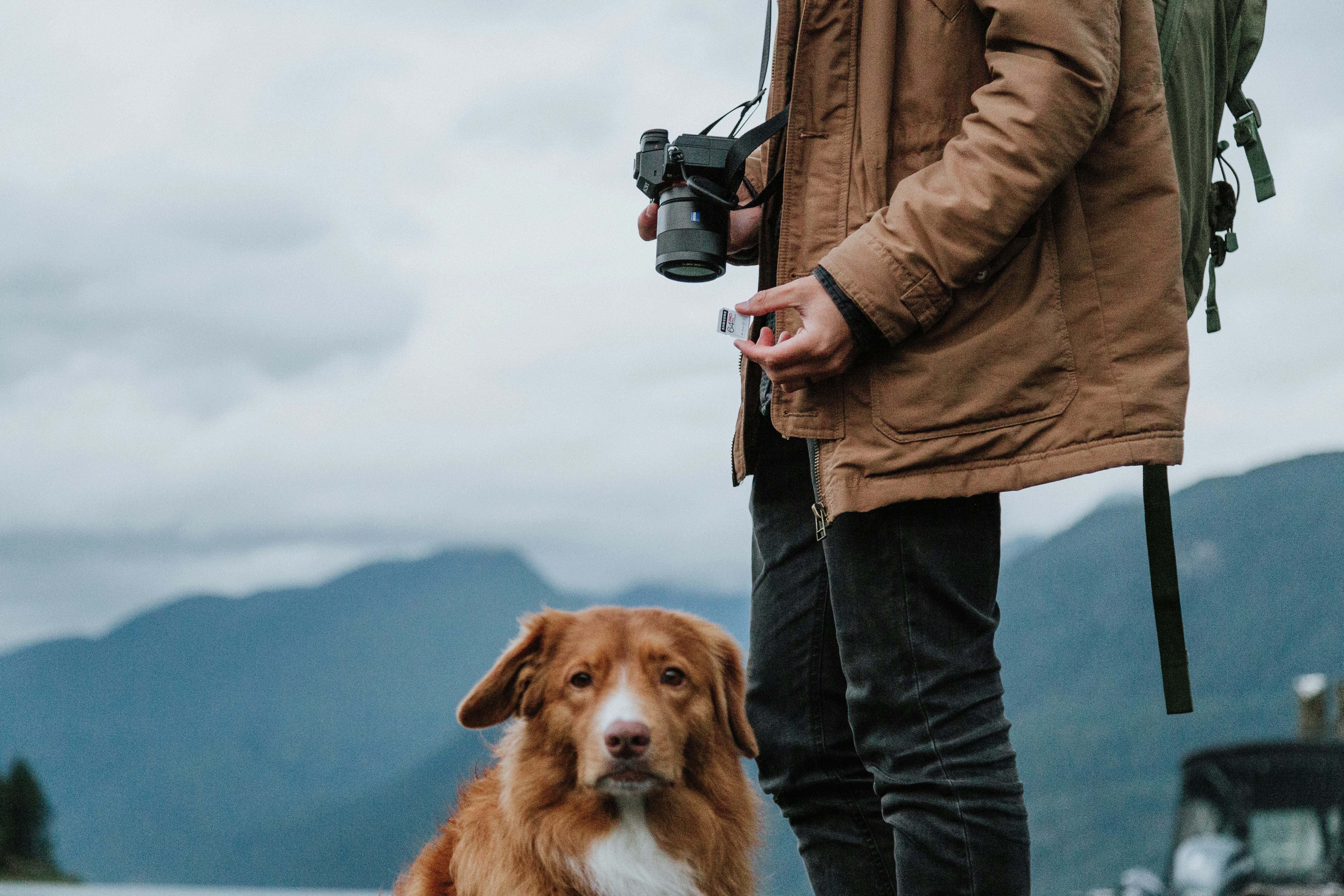 man in brown jacket and blue denim jeans holding black dslr camera