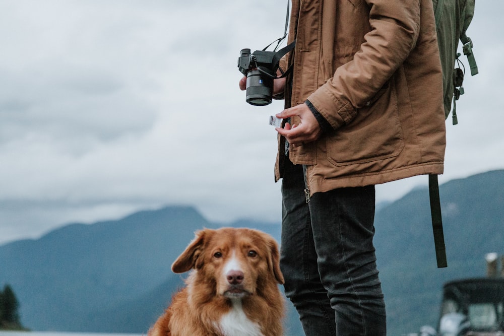 man in brown jacket and blue denim jeans holding black dslr camera