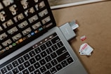 A laptop keyboard with a touch bar positioned next to a memory card adapter, into which a Samsung memory card is inserted. Several other microSD cards are scattered on the wooden surface nearby, each labeled with storage capacities.