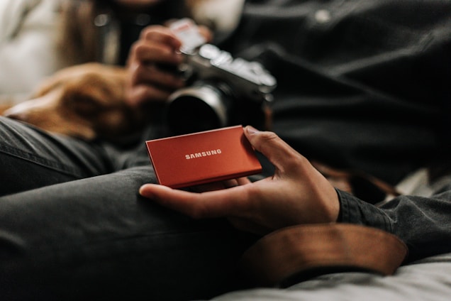 A person holds a small red Samsung external hard drive, with another hand in the background holding a camera. The setting appears to be casual, possibly on a sofa or bed with a relaxed atmosphere.