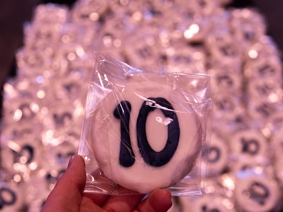 A hand is holding a cookie with the number 10 on it, covered in white icing and individually wrapped in clear plastic. In the background, there are multiple similar cookies scattered, all with numbers in a similar packaging.
