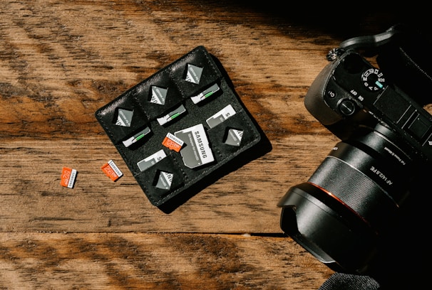 A wooden surface with a camera and a storage pouch containing memory cards. Several SD cards are arranged neatly in the pouch, with a few placed on the table. The camera is positioned to the right, showing part of its lens and control dials.