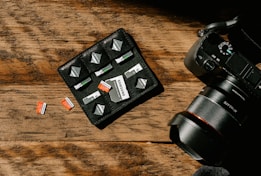 A wooden surface with a camera and a storage pouch containing memory cards. Several SD cards are arranged neatly in the pouch, with a few placed on the table. The camera is positioned to the right, showing part of its lens and control dials.